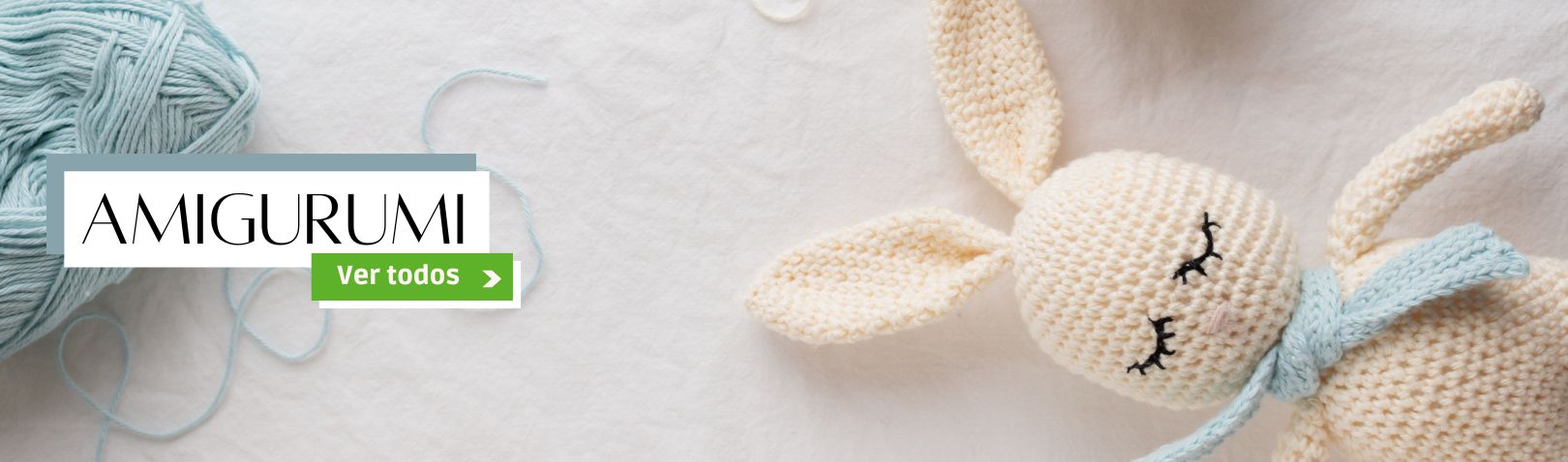 A imagem mostra uma propaganda de produtos para amigurumi. No lado esquerdo, há um novelo de linha azul claro e o texto "Amigurumi", com um botão verde escrito "Ver todos". No lado direito, aparece um boneco de crochê em formato de coelhinho, feito com linha bege e cachecol azul, deitado sobre um fundo branco.